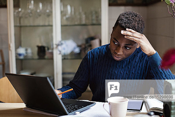 Besorgter Teenager schaut auf den Laptop  während er auf dem Tisch Hausaufgaben macht
