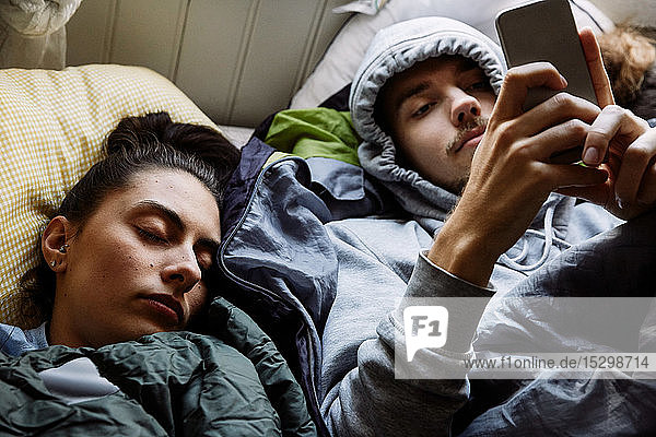 Schrägansicht eines jungen Mannes  der ein Mobiltelefon benutzt  während er bei einem Freund zu Hause im Bett liegt