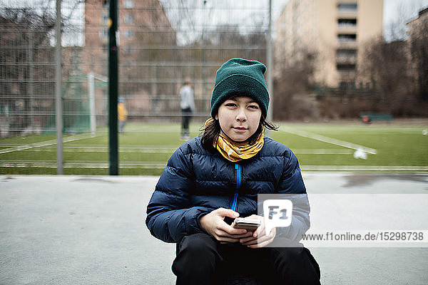 Porträt eines Jungen mit Mobiltelefon in warmer Kleidung  der in der Stadt vor einem Fussballfeld sitzt