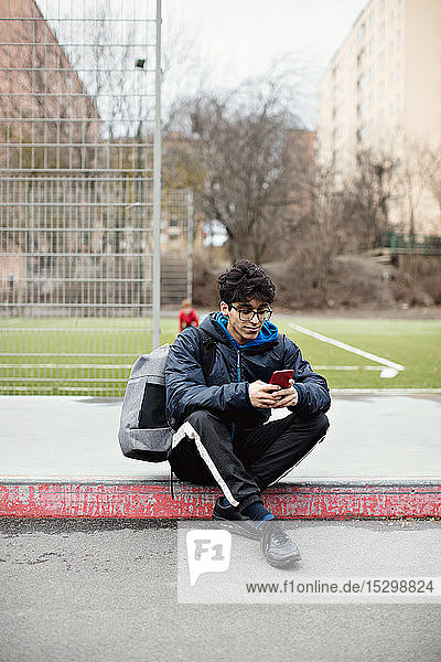 Ein junger Mann in voller Länge benutzt ein Mobiltelefon  während er im Winter auf dem Bürgersteig vor einem Fußballfeld sitzt