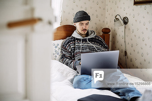 Junger Mann benutzt Laptop  während er entspannt auf dem Bett liegt und durch die Tür zu Hause gesehen wird