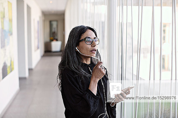 Geschäftsfrau spricht mit einem Smartphone  während sie im Büro am Fenster im Flur steht
