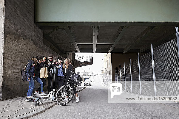 Männliche und weibliche Freunde im Teenageralter mit Roller und Fahrrad auf dem Bürgersteig unter der Brücke