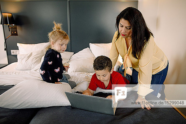 Mutter und Mädchen mit einem Jungen  der einen Laptop am Bett im Hotelzimmer benutzt