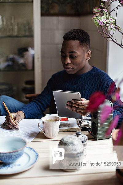 Teenager hält digitales Tablett in der Hand  während er zu Hause bei den Hausaufgaben auf ein Buch schreibt