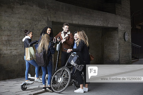 Lächelnde männliche und weibliche Freunde im Teenageralter mit Roller und Fahrrad auf dem Fußweg