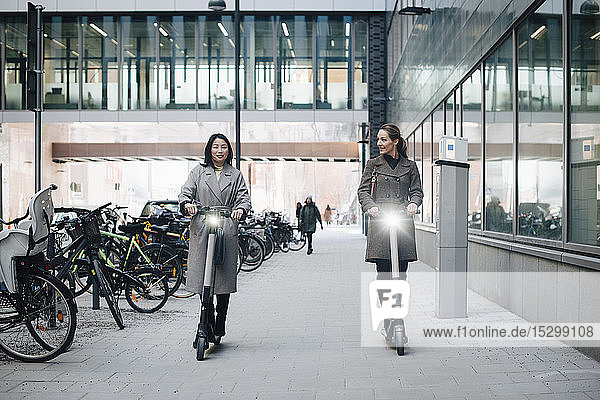 Selbstbewusste Kolleginnen auf Elektro-Schieberoller in voller Länge auf einem Fußweg durch ein Gebäude in der Stadt
