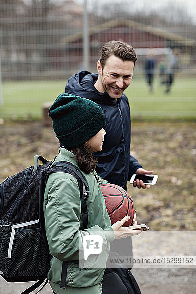 Lächelnder Vater und Sohn unterhalten sich beim Gehen auf der Straße nach dem Basketballtraining im Winter