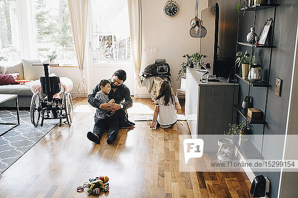 Schrägaufnahme eines Vaters  der einen autistischen Sohn hält  während die Tochter zu Hause auf dem Boden sitzt