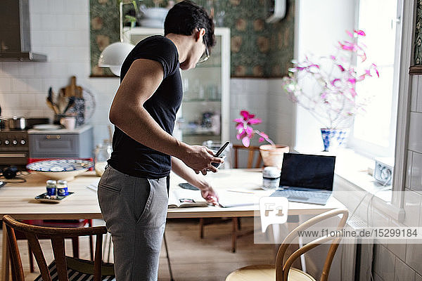 Seitenansicht eines jungen Mannes  der zu Hause stehend ein Buch vom Tisch nimmt