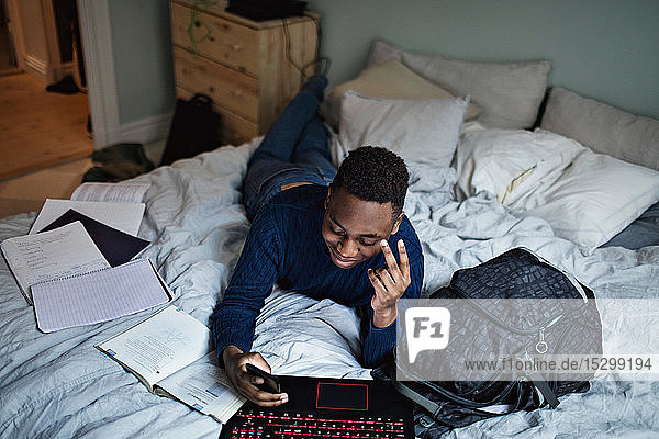Smiling teenage boy using social media while doing homework on bed at home