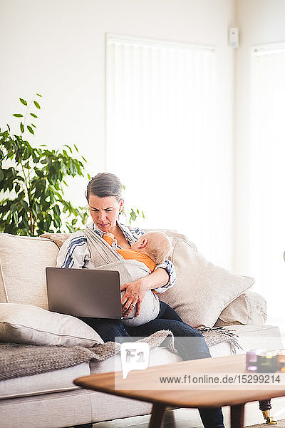 Multi-tasking mother using laptop while caring baby girl on sofa at home office