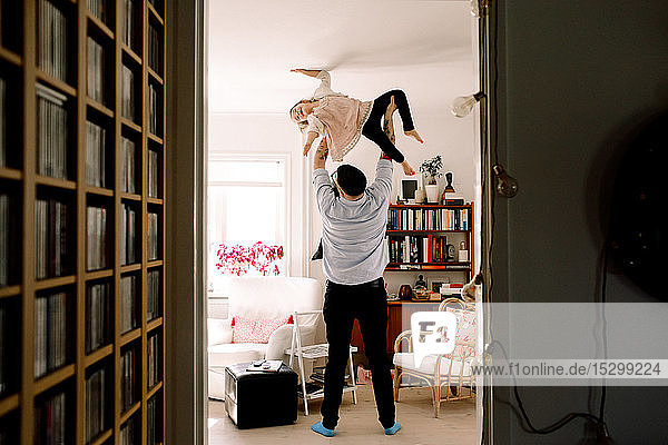 Verspielter Vater in voller Länge  der seine Tochter hochhebt  während er zu Hause im Wohnzimmer steht