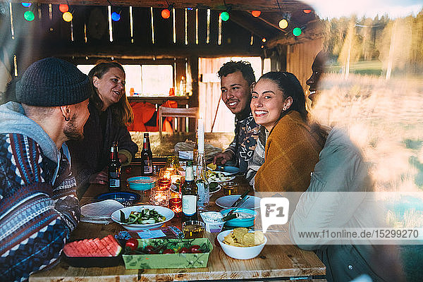 Männliche und weibliche Freunde unterhalten sich während des Essens auf dem Tisch in einer durch das Fenster gesehenen Hütte