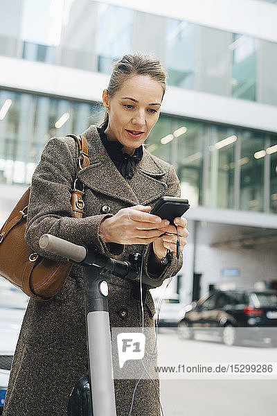 Selbstbewusste Geschäftsfrau benutzt ein Smartphone  während sie mit einem elektrischen Roller in der Stadt steht