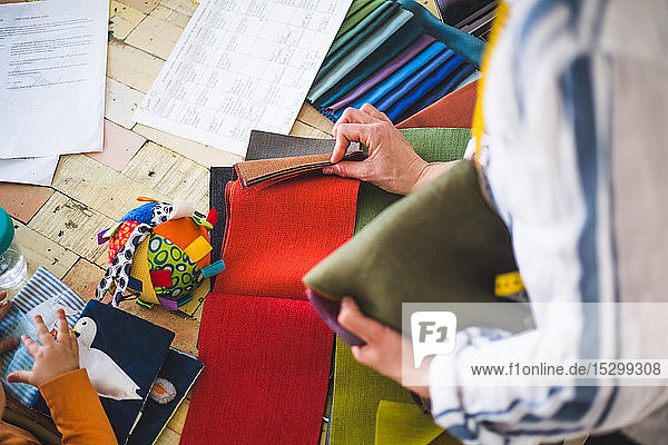 Hochwinkelansicht eines Modedesigners  der aus einem bunten Muster Stoff auswählt  während ein kleines Mädchen mit einem Stoffbild spielt