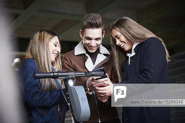 Glückliche männliche und weibliche Teenager-Freunde mit Elektroschubroller schauen auf das Smartphone unter der Brücke