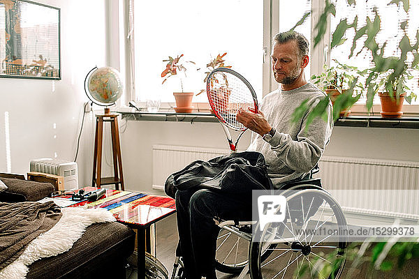 Nachdenklicher  reifer Mann hält Tennisschläger  während er zu Hause im Rollstuhl sitzt