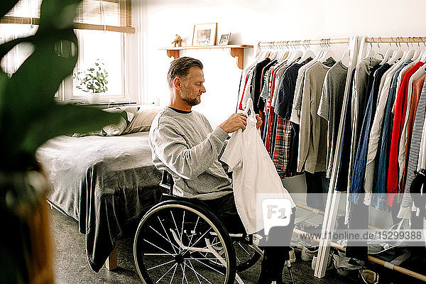 Seitenansicht eines behinderten reifen Mannes  der ein weißes T-Shirt hält  während er zu Hause am Kleiderständer im Rollstuhl sitzt