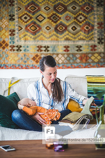 Working mother choosing fabric from swatch while breastfeeding baby girl in living room