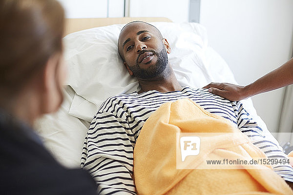 Junger Patient schaut Freund an  während er auf der Station im Krankenhaus auf dem Bett liegt