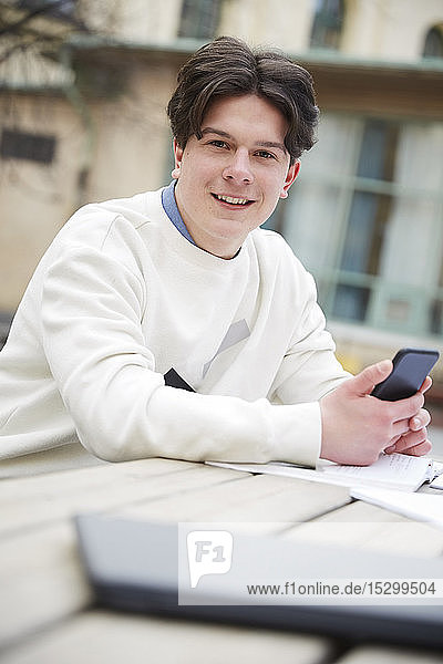 Portrait of confident teenage boy holding mobile phone while sitting at table in schoolyard
