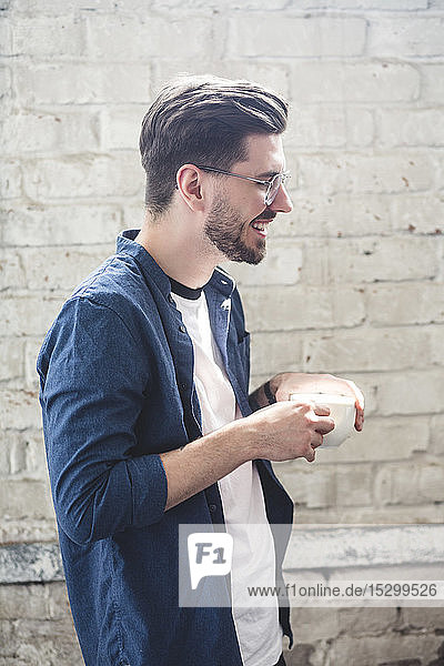 Seitenansicht eines bärtigen lächelnden Computerprogrammierers mit Kaffeetasse in der Hand  während er im Büro steht
