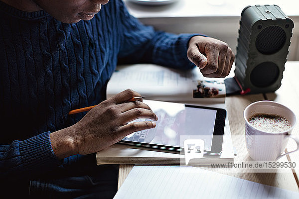 Mittelsektion eines Teenagers  der bei den Hausaufgaben ein digitales Tablett mit einer Kaffeetasse auf dem Tisch benutzt