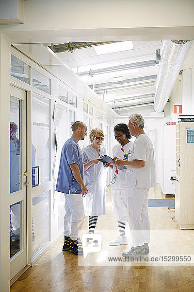 Mediziner diskutieren im Krankenhauskorridor stehend über ein digitales Tablett in voller Länge