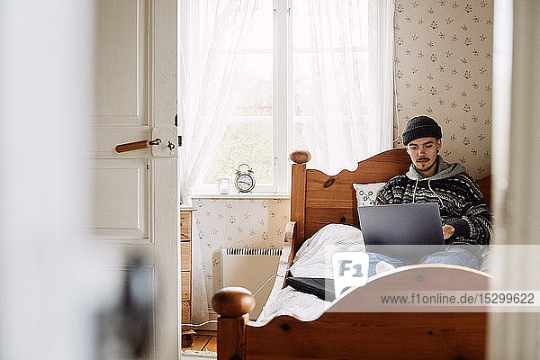 Junger Mann benutzt Laptop  während er zu Hause auf dem Bett sitzt und durch die Tür gesehen wird