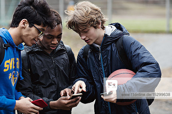 Friends looking in teenage boy's phone while standing with ball on playing field