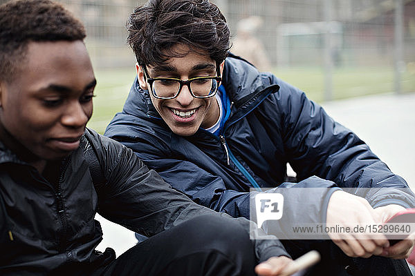 Lächelnder junger Mann  der das Handy eines Freundes gegen ein Spielfeld in der Stadt im Winter betrachtet