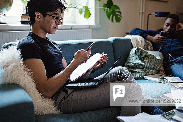 Young man studying while friend using social media on sofa at home