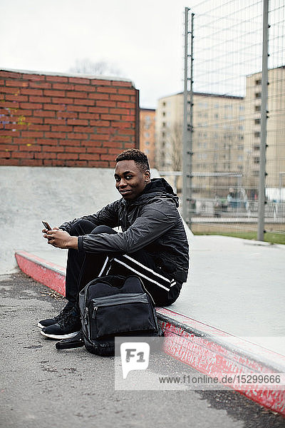 Side view portrait of smiling teenage boy using smart phone while sitting on sidewalk in city during winter