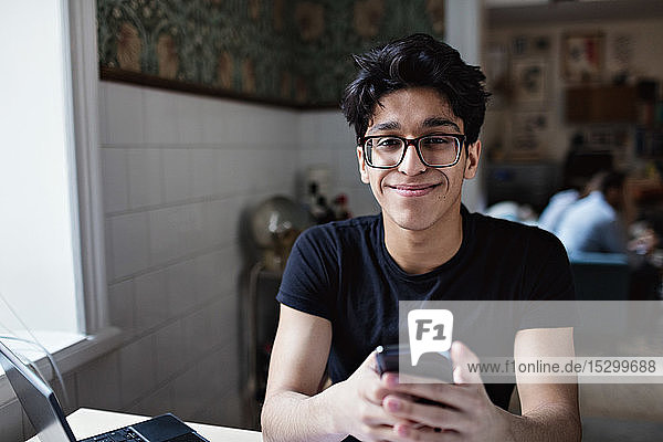 Porträt eines lächelnden jungen Mannes  der während des Studiums zu Hause soziale Medien auf seinem Mobiltelefon nutzt