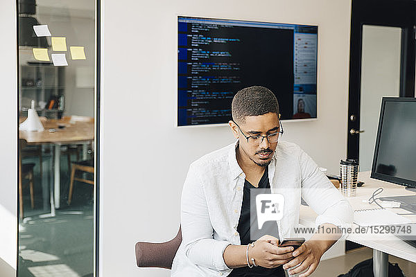 Männlicher Computerhacker  der ein Smartphone benutzt  während er am Schreibtisch im Kreativbüro sitzt