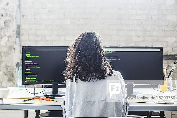 Rear view of female computer hacker coding at desk in creative office