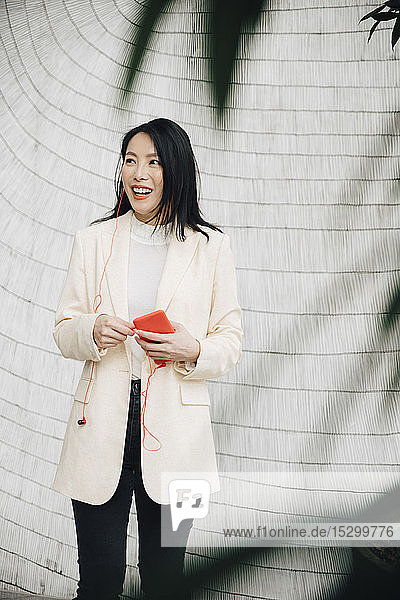 Lächelnde Geschäftsfrau hält Smartphone und Kopfhörer im Büro an die Wand