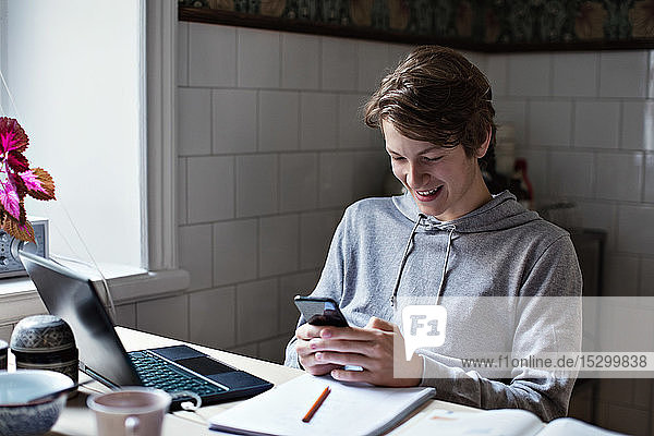 Lächelnder  Social-Media-süchtiger Teenager  der sein Handy benutzt  während er am Tisch Hausaufgaben macht