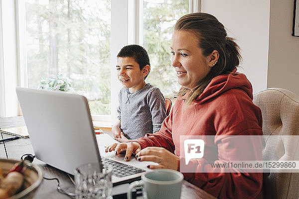 Mutter benutzt Laptop auf dem Tisch  während sie mit ihrem autistischen Sohn zu Hause sitzt