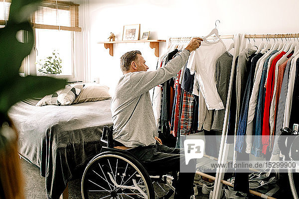 Seitenansicht eines behinderten reifen Mannes  der sein weißes T-Shirt zu Hause an einem Kleiderständer aufhängt