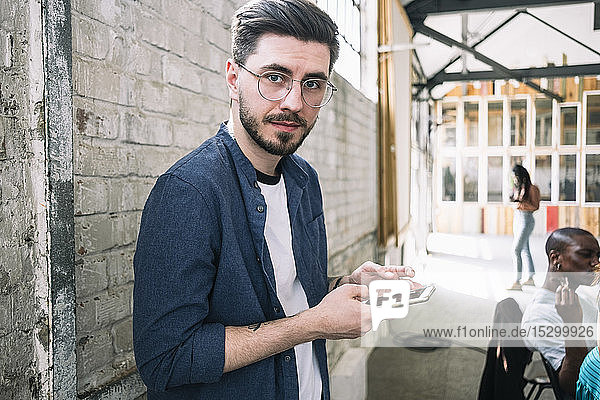 Portrait of confident young businessman standing with smart phone against brick wall at creative workplace