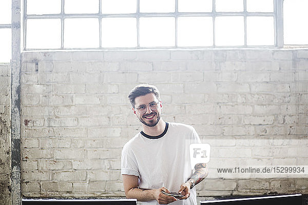 Porträt eines selbstbewusst lächelnden jungen Geschäftsmannes  der am Arbeitsplatz mit seinem Smartphone an einer Ziegelmauer steht