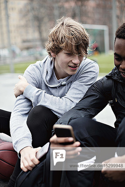 Teenager  der nach dem Basketballtraining auf dem Bürgersteig sitzend auf das Handy eines Freundes schaut