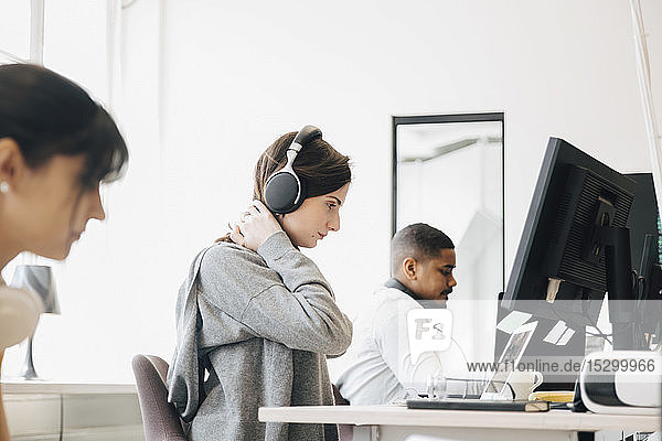 Focused female programmer with headphones using laptop on desk while sitting with coworkers in office