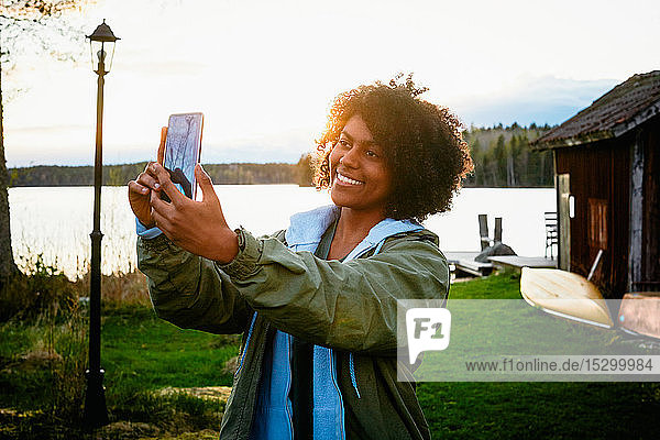 Lächelnde junge Frau  die bei Sonnenuntergang am See steht und sich mit einem Smartphone selbstständig macht