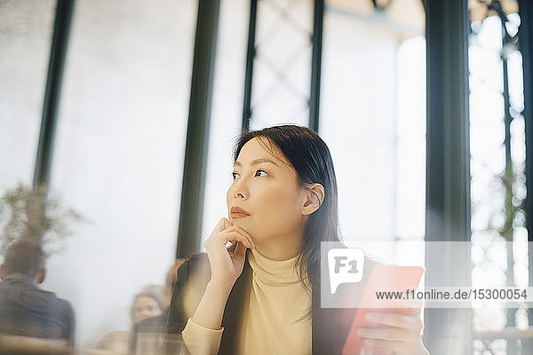 Nachdenkliche Geschäftsfrau hält Smartphone in der Hand  während sie im Büro auf die Cafeteria schaut