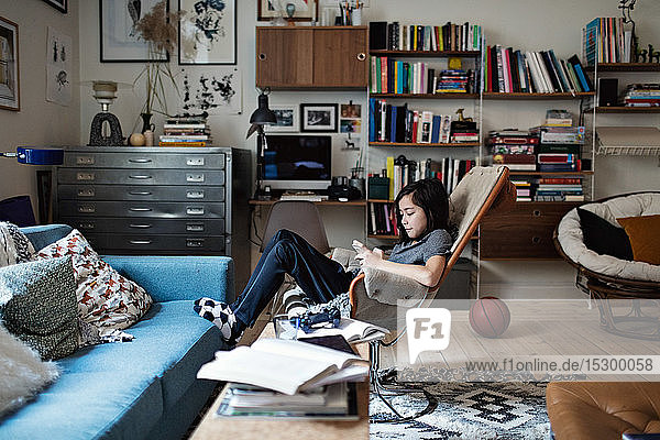 Junge in voller Länge  der ein Smartphone benutzt  während er zu Hause im Wohnzimmer sitzt