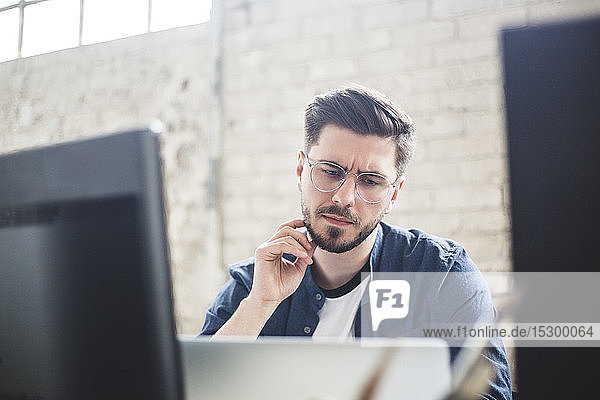 Nachdenklicher junger IT-Experte berührt Bart  während er im Büro auf den Laptop schaut