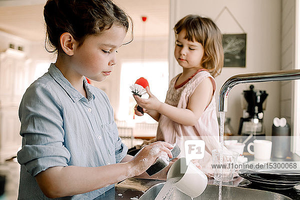 Mädchen wäscht Tasse  während die Schwester mit der Reinigungsbürste an der Küchentheke spielt